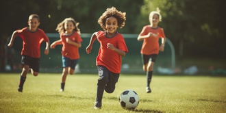  Das Foto zeigt vier Kinder frontal und lächelnd beim Fußballtraining. Das Training findet draußen auf einem Rasen statt. Im Vordergrund befindet sich ein Fußball. Im Hintergrund befindet sich ein Fußballtor.